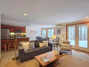 A modern living room with a grey sofa, a wooden coffee table, and a dining area with a chandelier. - Spacious 2-BR Ski-in, Ski-out Jay Peak Village Condo, right on trail. A/C incl! (Jay)