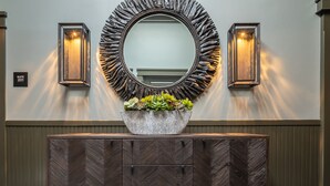 A mirror with a textured frame, flanked by wall-mounted lights, above a wooden cabinet with a plant.