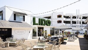 Zona de estar al aire libre frente al agua, con sillas y mesas de mimbre, un edificio blanco con el letrero 'LIDO MARINA VILLAGE' y luces colgantes.