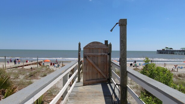A wooden boardwalk leading to a beach with a wooden gate, a pier in the background, and people enjoying the beach.