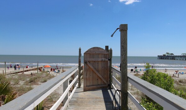 Ein Holzsteg führt zu einem Strand, an dem ein Holztor steht. Im Hintergrund ist ein Steg zu sehen, und Menschen entspannen sich am Strand.
