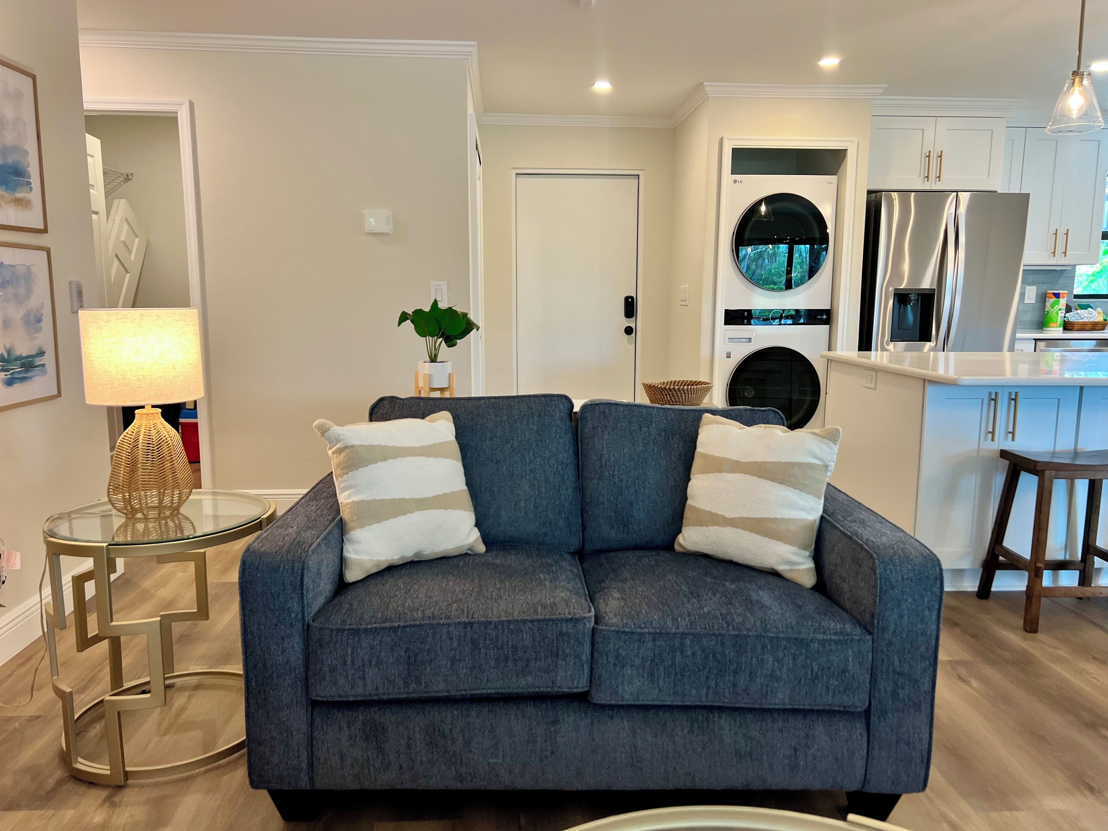 A modern living room with a grey sofa, a glass coffee table, and a lamp. A kitchen with white cabinets and stainless steel appliances is visible in the background.