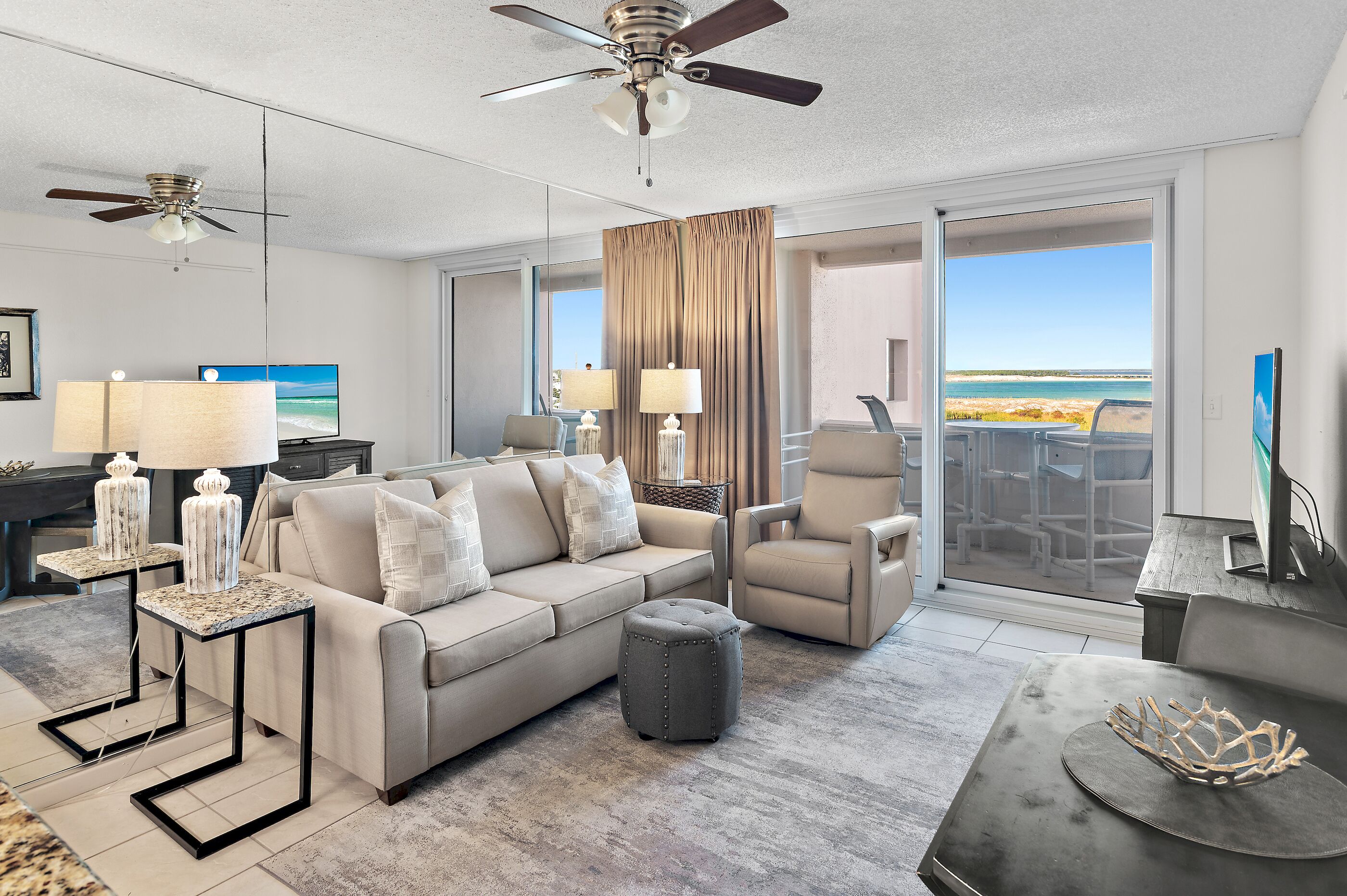 Ein modernes Wohnzimmer mit Sofa, Sesseln und Hocker, Deckenventilator und einem großen Fenster mit Blick auf den Strand.