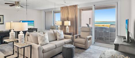 Ein modernes Wohnzimmer mit Sofa, Sesseln und Hocker, Deckenventilator und einem großen Fenster mit Blick auf den Strand.