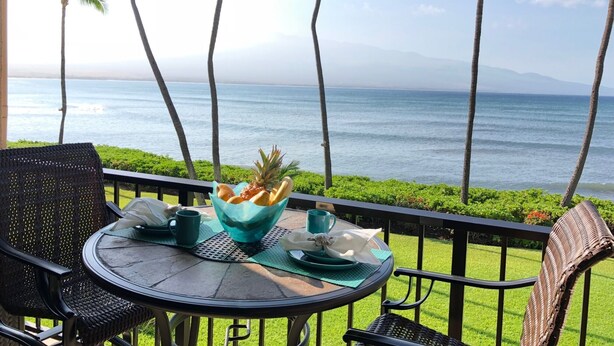 Une table avec un saladier de fruits, une tasse et une assiette, sur un balcon donnant sur l’océan, avec des palmiers en arrière-plan.