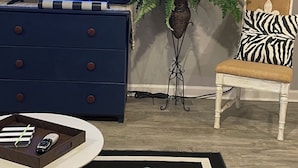 A living room with a television displaying a news program, a clock on the wall, a blue dresser, a chair with a zebra-patterned pillow, and a potted plant.