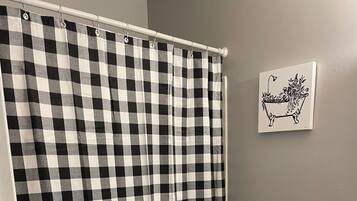 Un bagno con tenda da doccia a scacchi bianchi e neri, un water, una parete bianca con un quadro incorniciato e un cesto bianco contenente asciugamani.