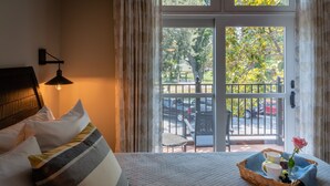 Habitación de hotel con cama, mesita de noche, lámpara, vista al exterior por una puerta de vidrio y balcón con una silla y una mesa.