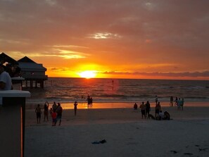 A beach scene with a sunset, people walking, and beach huts in the background.