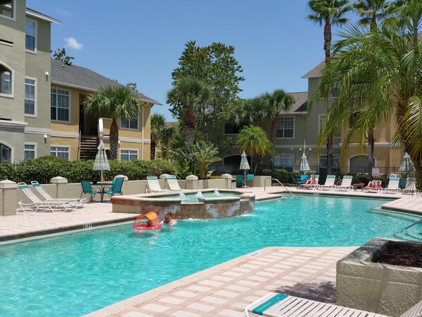 A pool area with lounge chairs, umbrellas, and a hot tub.