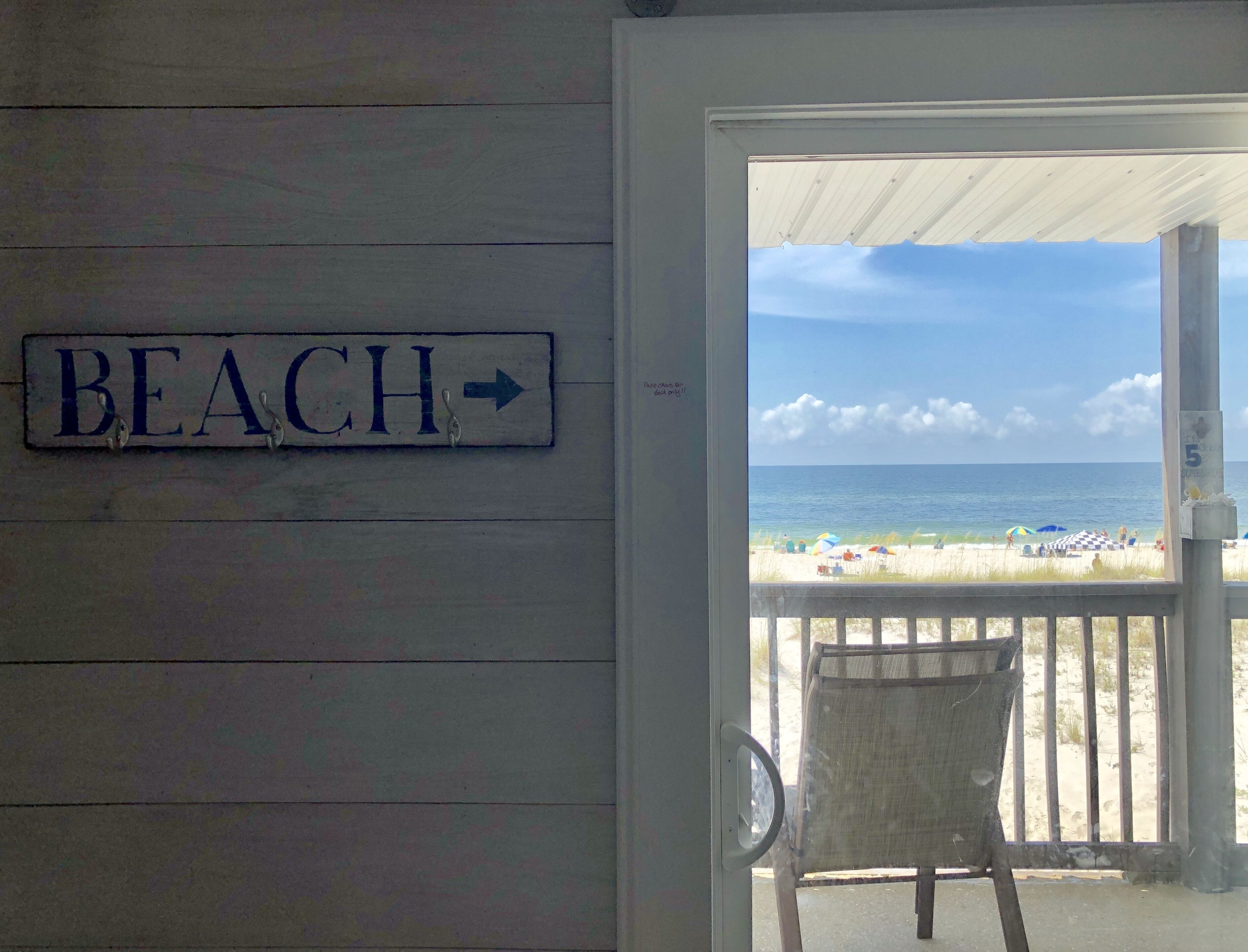Puerta de madera con un letrero que dice "BEACH" y vista de una playa con sombrillas y gente.