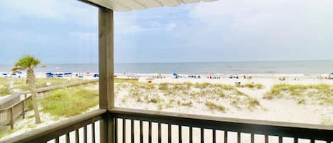 A beach view from a covered balcony with a railing, a palm tree, and beach umbrellas.