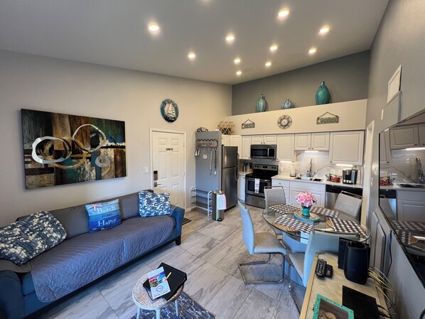 A modern kitchen with white cabinets, a black stove, and a microwave. There is a dining table with chairs and a small rug. A gray sofa with blue pillows is in the living area.