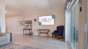 A spacious living room with a light-colored floor, a dark armchair, a coffee table, and a bookshelf. A painting is on the wall, and a sign reads "The Best Rentals". - Spacious Condo Close to Everything! -Pier Pleasure (Folly Beach)