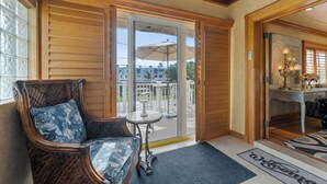 A wicker chair with a floral cushion, a small round table, and a welcome mat in a room with wooden paneling and a view through a glass door to an outdoor patio.