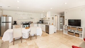 Una cocina moderna con armarios blancos, un comedor con mesa dispuesta para cuatro personas y un televisor en un mueble blanco.