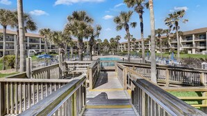 A wooden boardwalk leading to a pool area surrounded by palm trees and a multi-story building with balconies. - Ocean Villas 108: Top floor 3 bedroom condo in gated complex - 2 pools, beach (St. Augustine Beach)