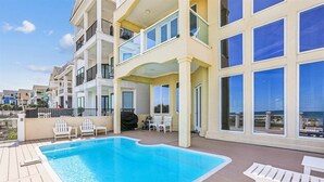 A multi-story building with a pool area featuring a wooden deck, white lounge chairs, and large windows offering ocean views. - 12 Collier Beach, Oceanfront, Pool, Great for Large Groups, Free Golf, Oceanfront (Hilton Head Island)