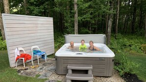 A hot tub in a garden with a wooden deck, stone pathway, and a white fence.
