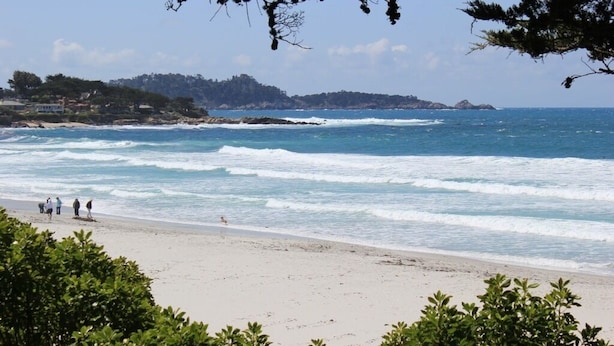 Une plage avec des vagues, quelques personnes et une côte lointaine parsemée de bâtiments.