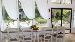 Un coin repas avec une table en bois et des chaises blanches, un panier de fruits et une bouteille de vin.