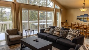 A cozy living room with a dark-colored sofa, a small coffee table, and a chair. There is a large window with curtains, a balcony visible outside, and a chandelier hanging from the ceiling.