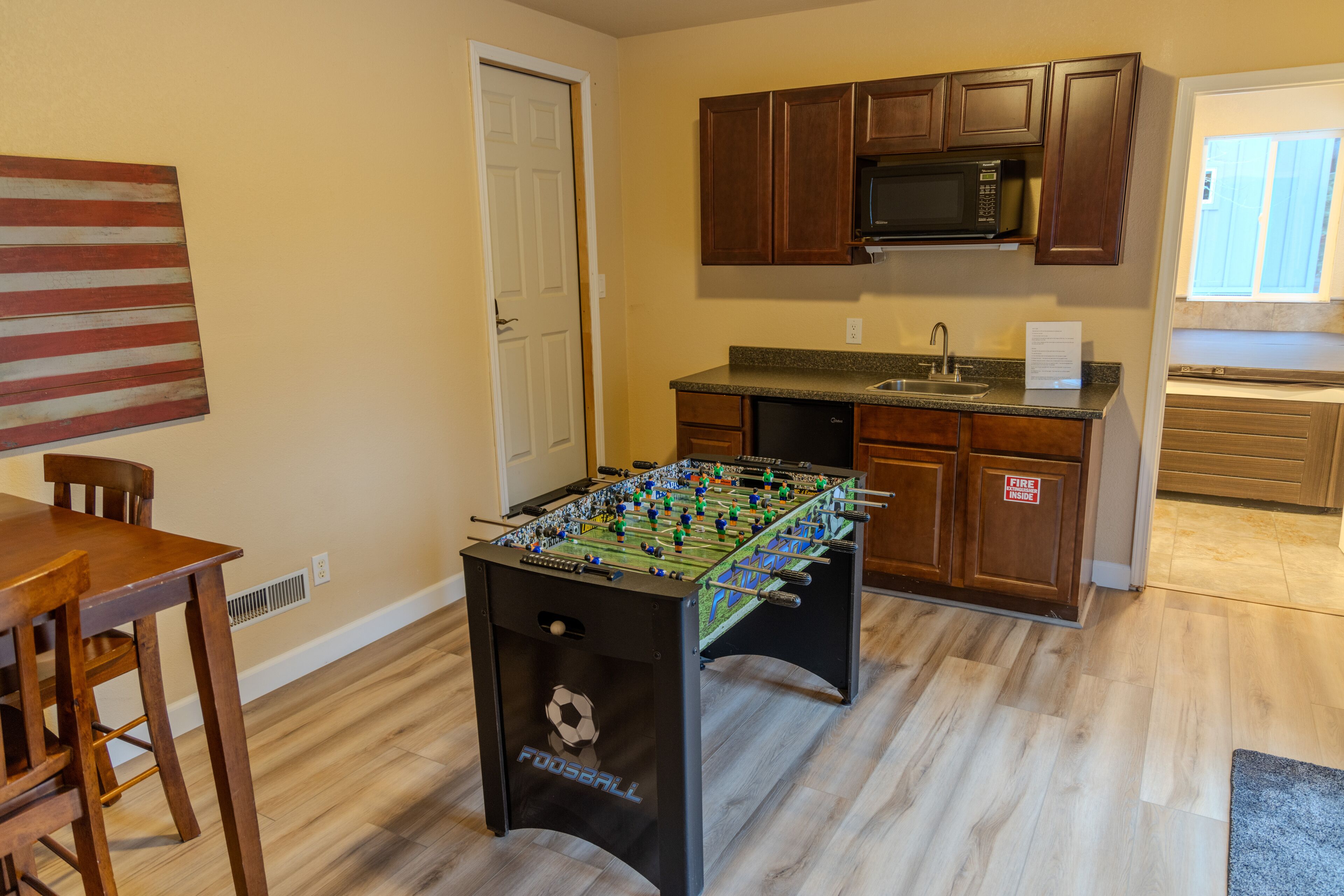 Una cocina con armarios de madera, mesa de futbolín, mesa de comedor con sillas y ventana con persianas.