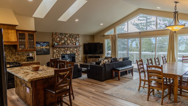 A spacious kitchen with a large island, wooden cabinets, and a stone wall. There is a dining area with a table and chairs, a sofa, and a TV.