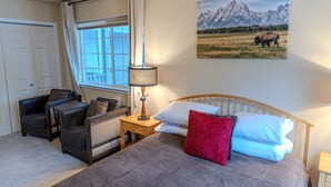 A bedroom with a bed, two armchairs, a nightstand with a lamp, a window with curtains, and a wall-mounted picture of a bison in a field.