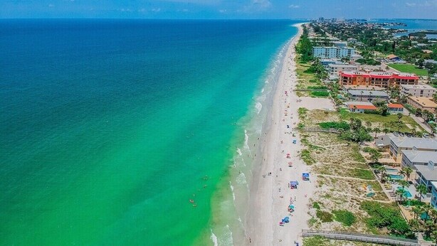 An aerial view of a beach with clear turquoise waters, a sandy shore, and buildings along the coastline. - 2-Story Beach-side Complex with Direct Beach Access, Book Your Beach Getaway! (Indian Rocks Beach)
