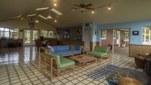 Un vaste espace de vie avec un canapé bleu, des fauteuils verts et une table basse en bois. On y trouve des ventilateurs de plafond, un coin cuisine à l’arrière-plan et un sol à motifs.