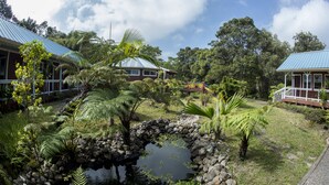 Un jardin tropical avec un étang, entouré d’une végétation luxuriante et bordé de deux bâtiments aux toits bleus.