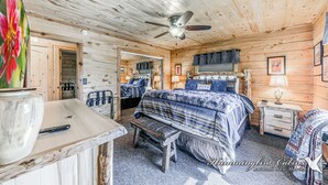 Interior de una cabaña con cama, escritorio, silla, ventilador de techo y ventana con cortinas.
