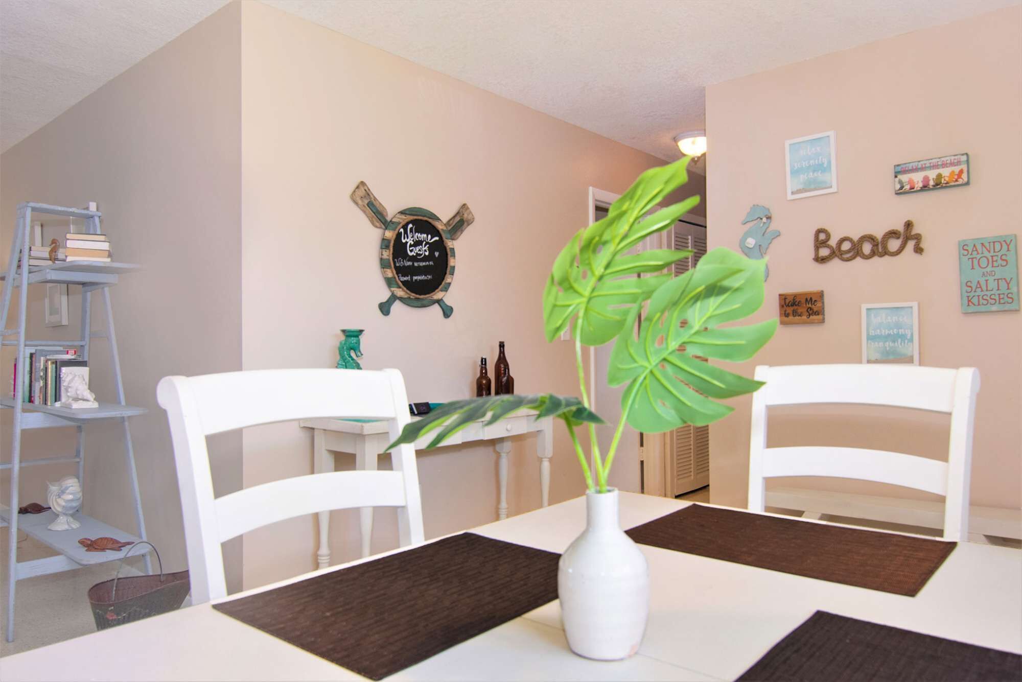 Un coin repas avec deux chaises blanches, une table blanche et un vase blanc contenant une plante. Sur la gauche, on aperçoit une bibliothèque remplie de livres et d’objets décoratifs. Le mur est orné de plusieurs tableaux encadrés et d’une enseigne indiquant le mot « Plage ».