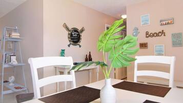 Un coin repas avec deux chaises blanches, une table blanche et un vase blanc contenant une plante. Sur la gauche, on aperçoit une bibliothèque remplie de livres et d’objets décoratifs. Le mur est orné de plusieurs tableaux encadrés et d’une enseigne indiquant le mot « Plage ».