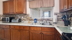 Una cocina con armarios de madera, microondas, tostadora, lavamanos y una ventana con cortina.