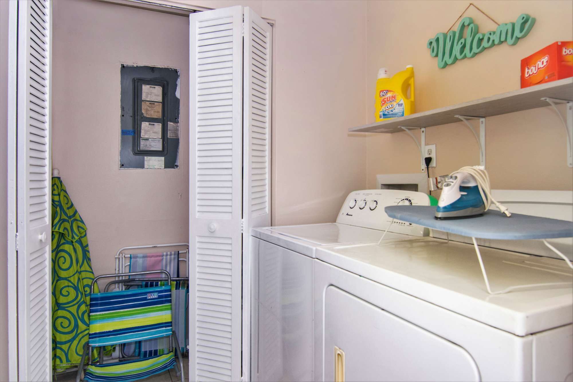A laundry area with a washing machine, an ironing board, and a 'Welcome' sign.