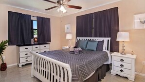 Un dormitorio con cama, mesitas de noche, ventilador de techo y ventana con cortinas.