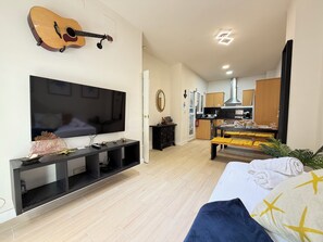 Un salon avec un téléviseur, une guitare, un coin repas avec des coussins jaunes et une cuisine à l’arrière-plan.