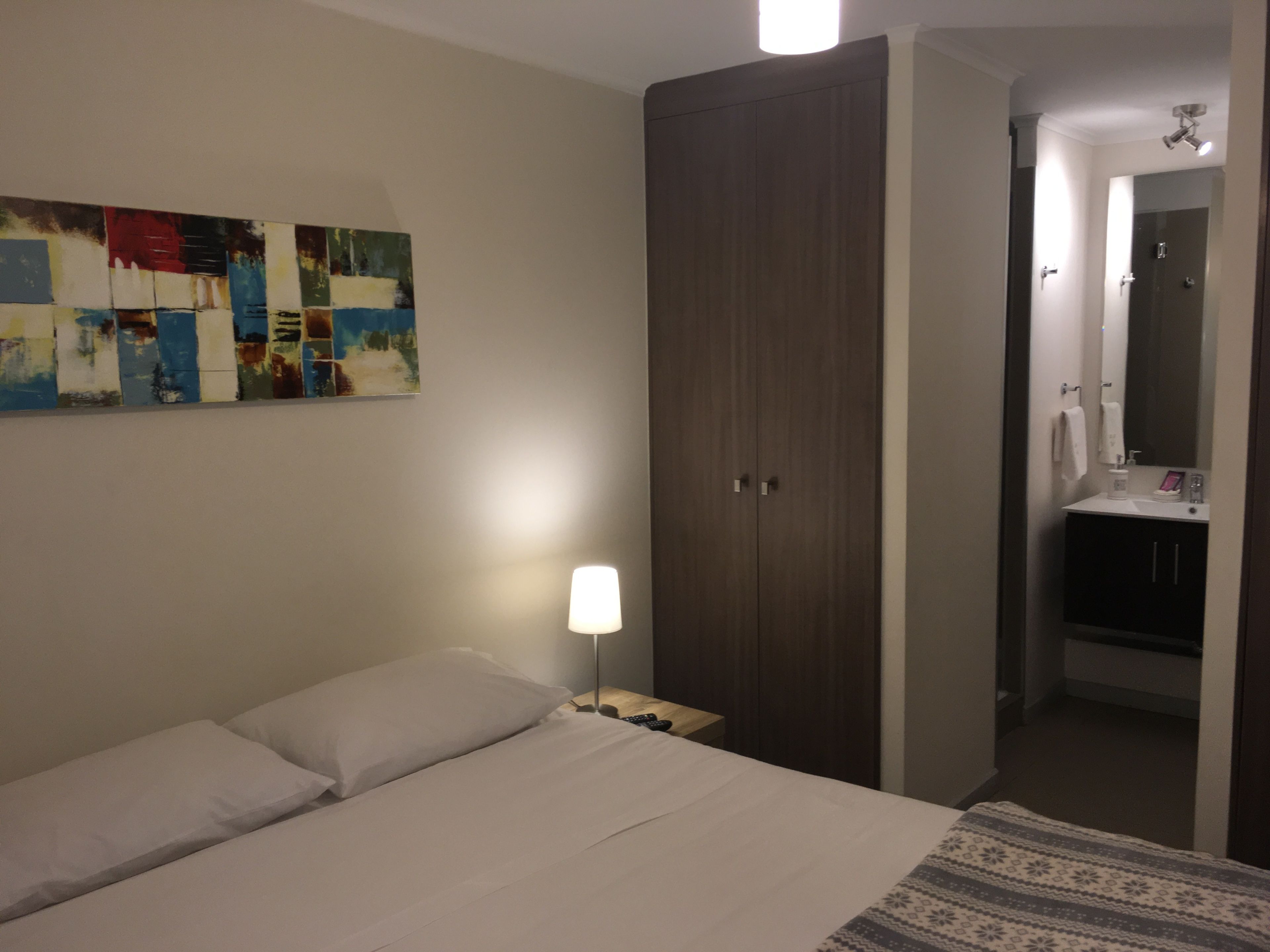 Una habitación de hotel con cama, mesita de noche, armario, baño con lavamanos y espejo, y una pared con un cuadro abstracto colorido.