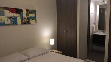 Una habitación de hotel con cama, mesita de noche, armario, baño con lavamanos y espejo, y una pared con un cuadro abstracto colorido.