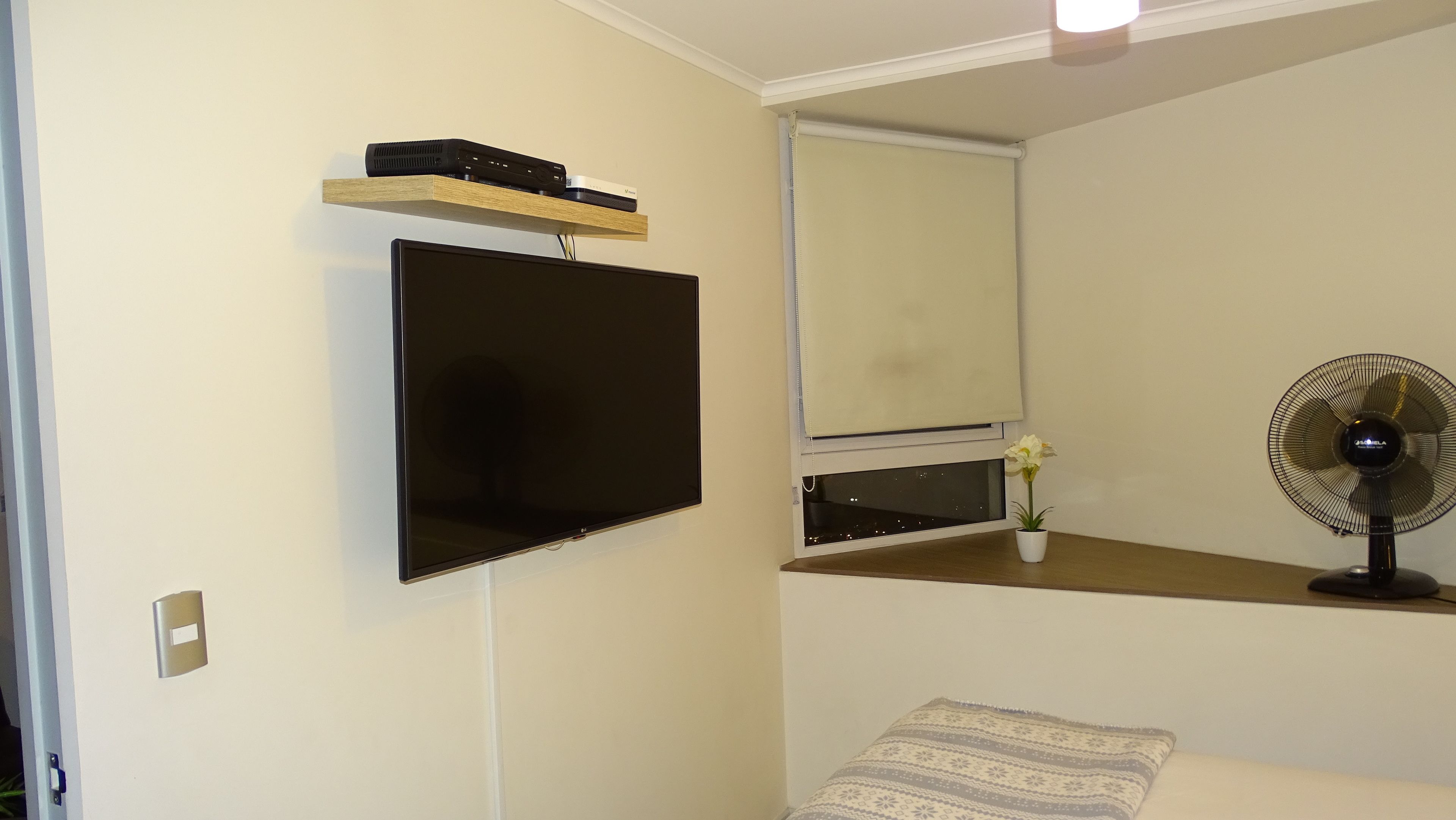 Una habitación con una televisión montada, un estante con dispositivos electrónicos, una ventana con persiana, un ventilador de pie y una mesita de noche con un cojín estampado.