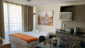 A hotel room with a bed, a desk, a chair, a dining table, a microwave, a kettle, and a painting of a cityscape. - Moneda Express Apart (Santiago)