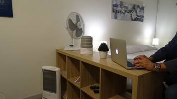 A hotel room with a wooden bedside table, a laptop, a fan, a small potted plant, a remote control, and a wall-mounted picture.