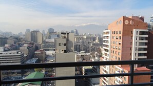 A cityscape with high-rise buildings, a mountain range in the background, and a view through a window with a metal frame. - Moneda Express Apart (Santiago)