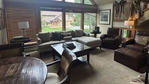 A spacious living room with a high ceiling, large windows, a sectional sofa, leather armchairs, a wooden coffee table, and a stone fireplace.
