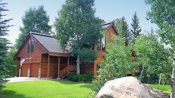 A wooden cabin with a porch, surrounded by lush greenery and a large rock in the foreground.