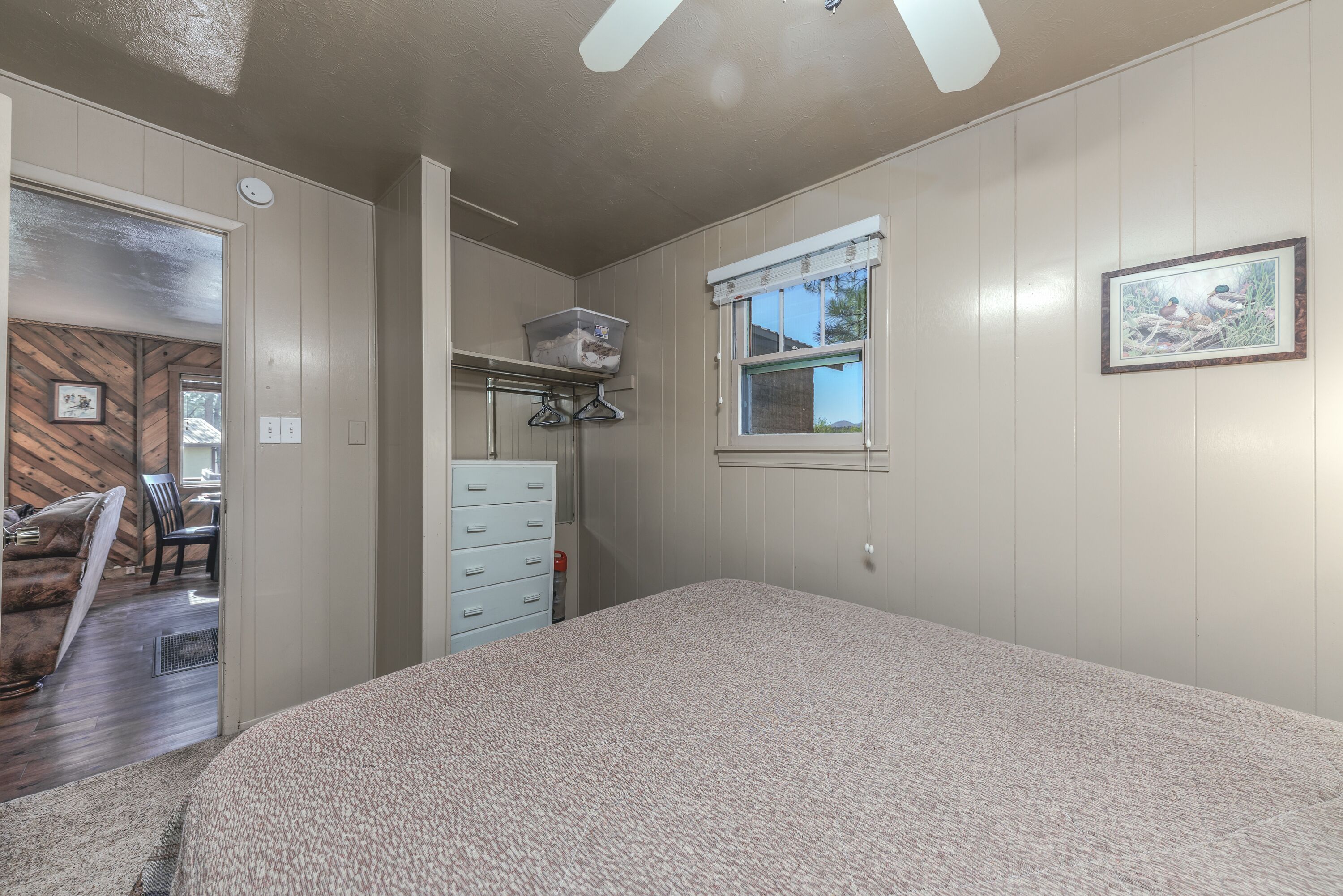 A bedroom with a large bed, a dresser, a window with blinds, a ceiling fan, and a wooden wall adjacent to the room.