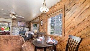 A wooden interior with a dining table set for two, a fireplace, and a television in the background. - Happy Days Inn | Your Happy Place in the Pines w/ Hot Tub,Views & Pet Friendly (Ruidoso)