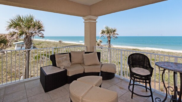 A covered patio with a wicker sofa, a round ottoman, a wicker chair, and a small table. The patio overlooks a beach with palm trees and a clear blue sky. - Gorgeous Oceanfront 3Bedroom Away From The Hustle and Bustle (St Augustine)